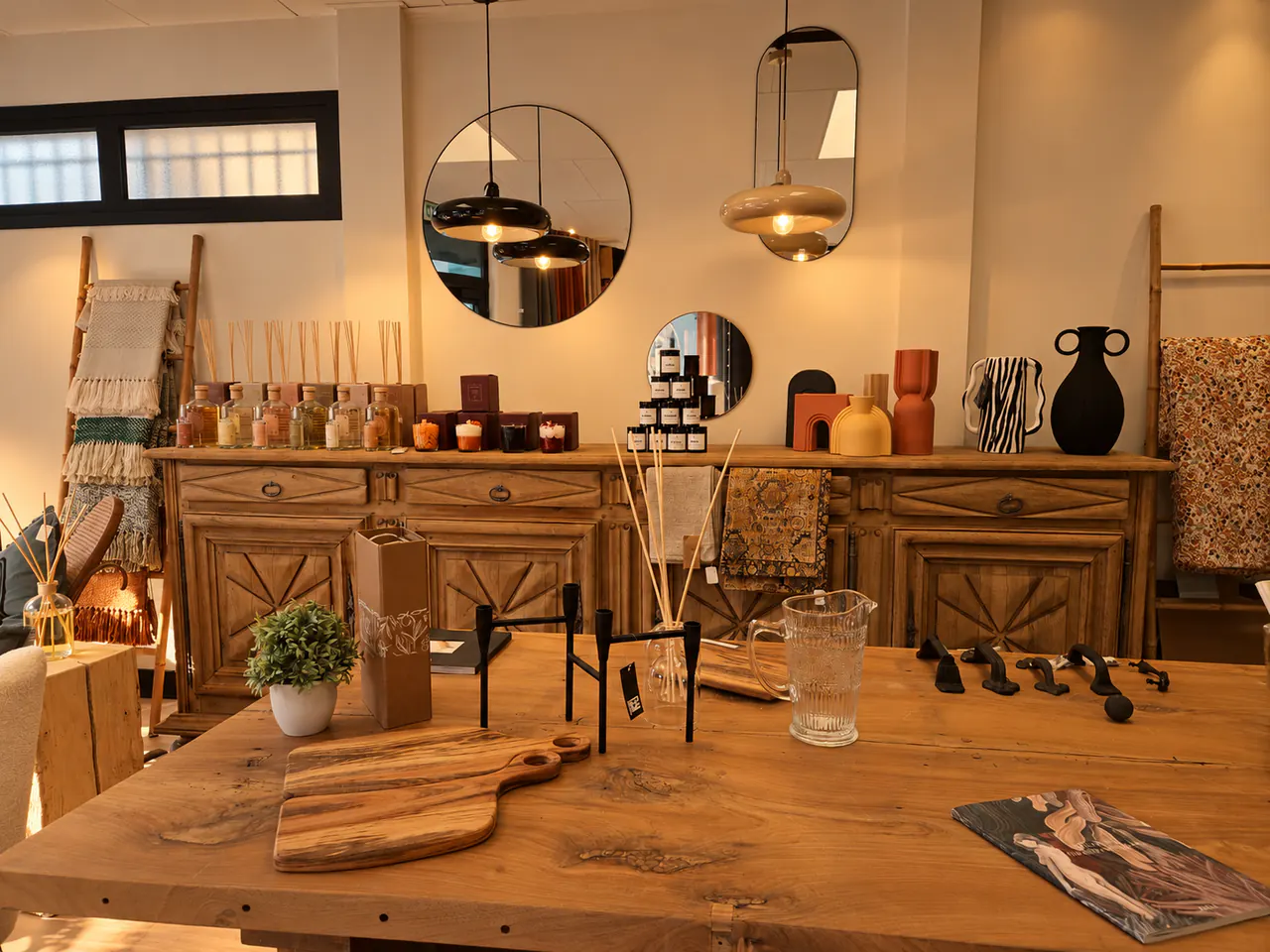 Le buffet sculpté chargé de bougies, diffuseurs et vases, sous des miroirs et des suspensions, devant une table de ferme en chêne